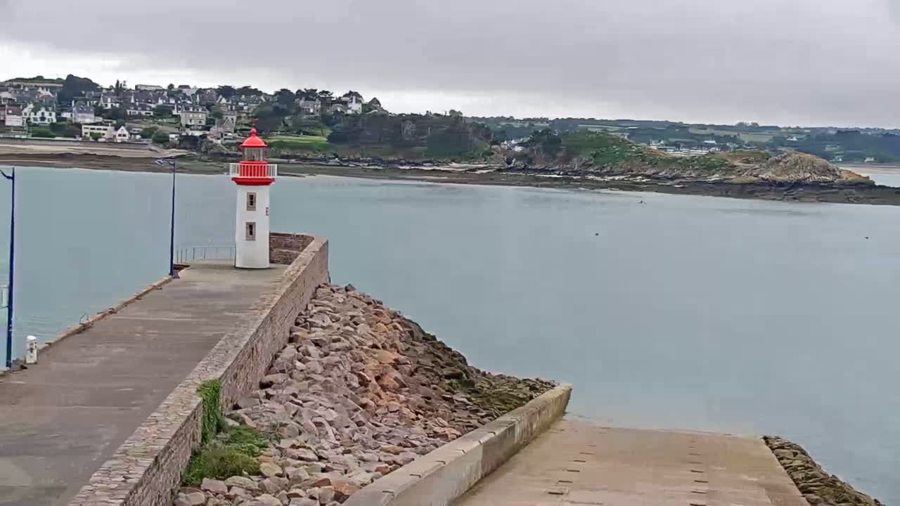 A red and white lighthouse stands on a stone jetty overlooking a calm, gray body of water, with a town and small island visible in the distance under an overcast sky.