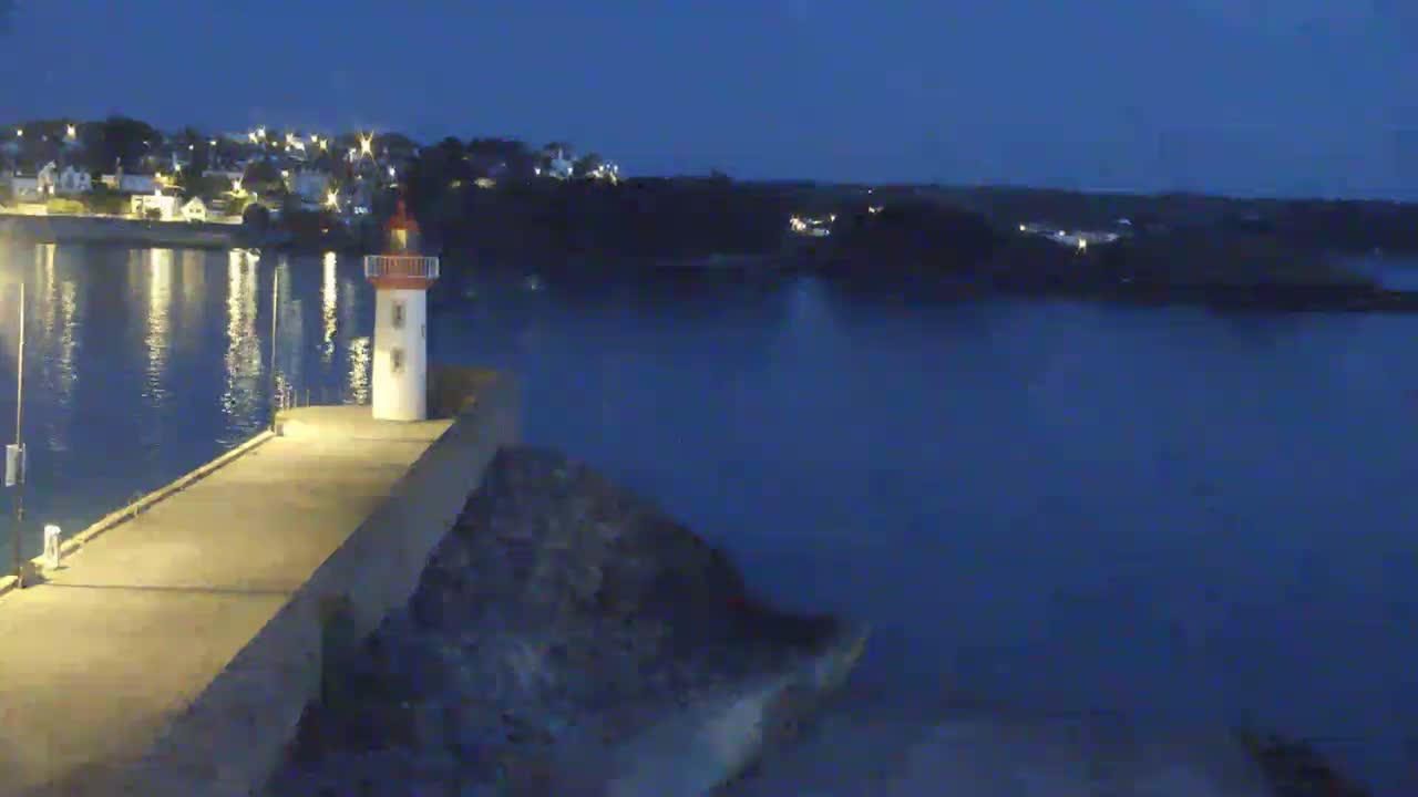 A small lighthouse stands on a pier at night, overlooking calm, dark water and a town with lights in the distance.
