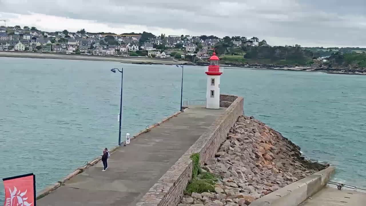 A person walks along a paved path on a stone breakwater leading to a lighthouse, overlooking a calm sea and a town under a cloudy sky.