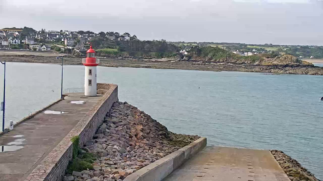A red and white lighthouse stands on a stone breakwater overlooking a calm body of water and a distant coastline under an overcast sky.