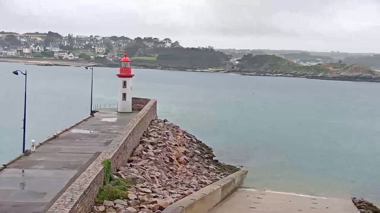 A red and white lighthouse stands on a stone breakwater overlooking a calm, gray-toned body of water and a distant town under an overcast sky.