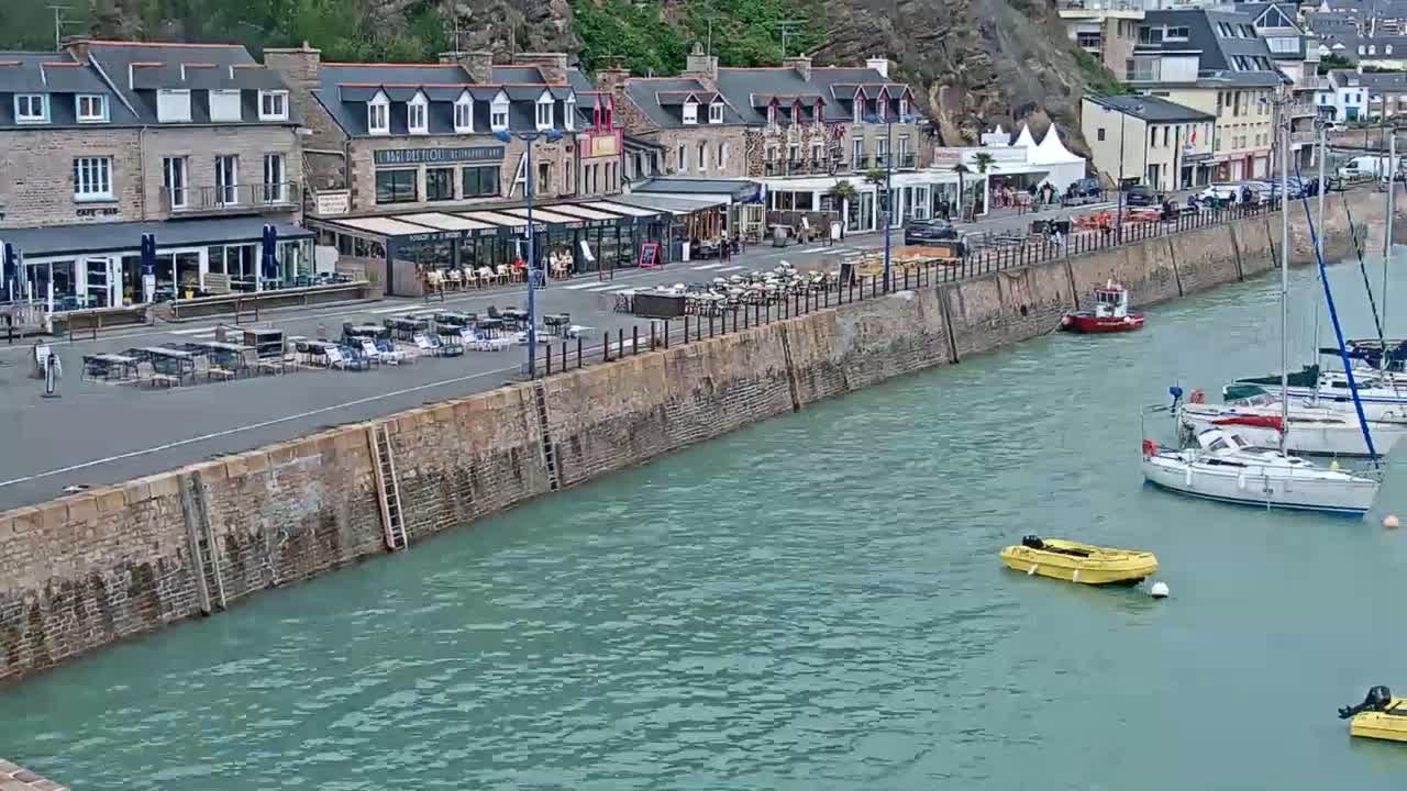 A harbor scene with calm, teal water shows several sailboats moored alongside a stone wall, behind which are restaurants with outdoor seating and buildings on a hillside.