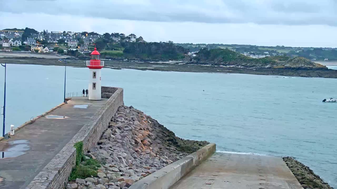 A red and white lighthouse stands on a stone pier overlooking a calm body of water under an overcast sky, with a small town and distant coastline visible in the background.