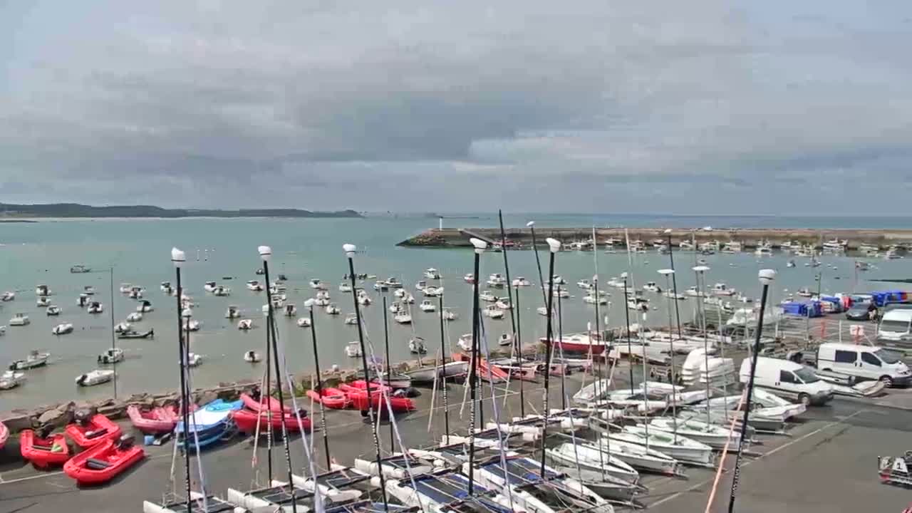 Numerous sailboats and small motorboats are docked in a harbor on a cloudy day, with additional boats anchored in the calm water beyond.