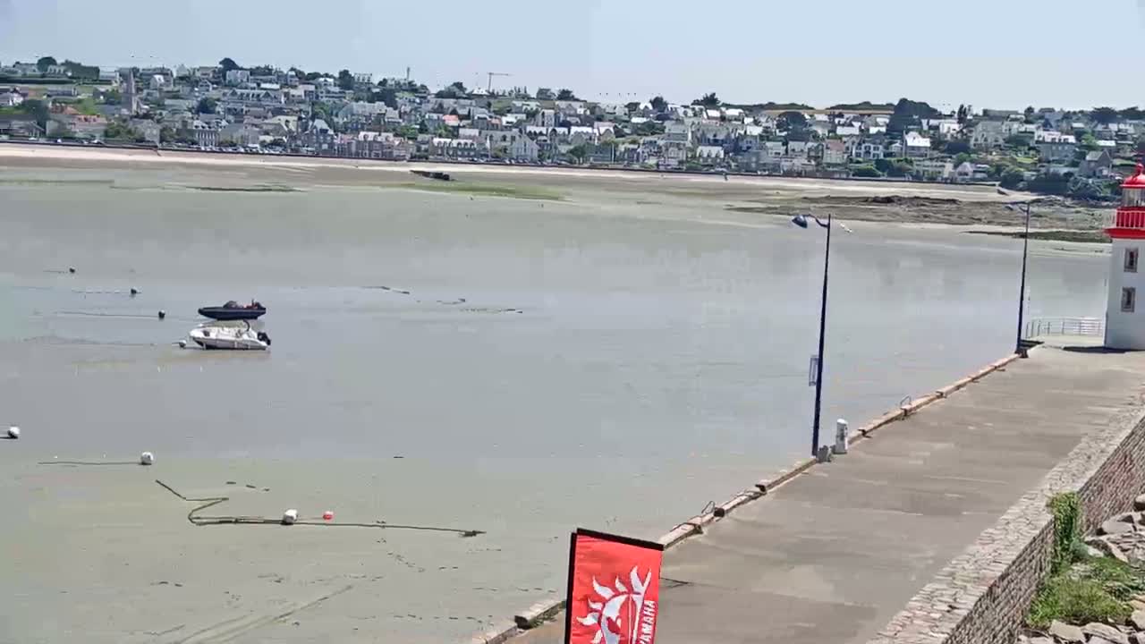 A coastal town is visible across a calm body of water, partially exposed at low tide, with two boats and a lighthouse on a nearby pier.