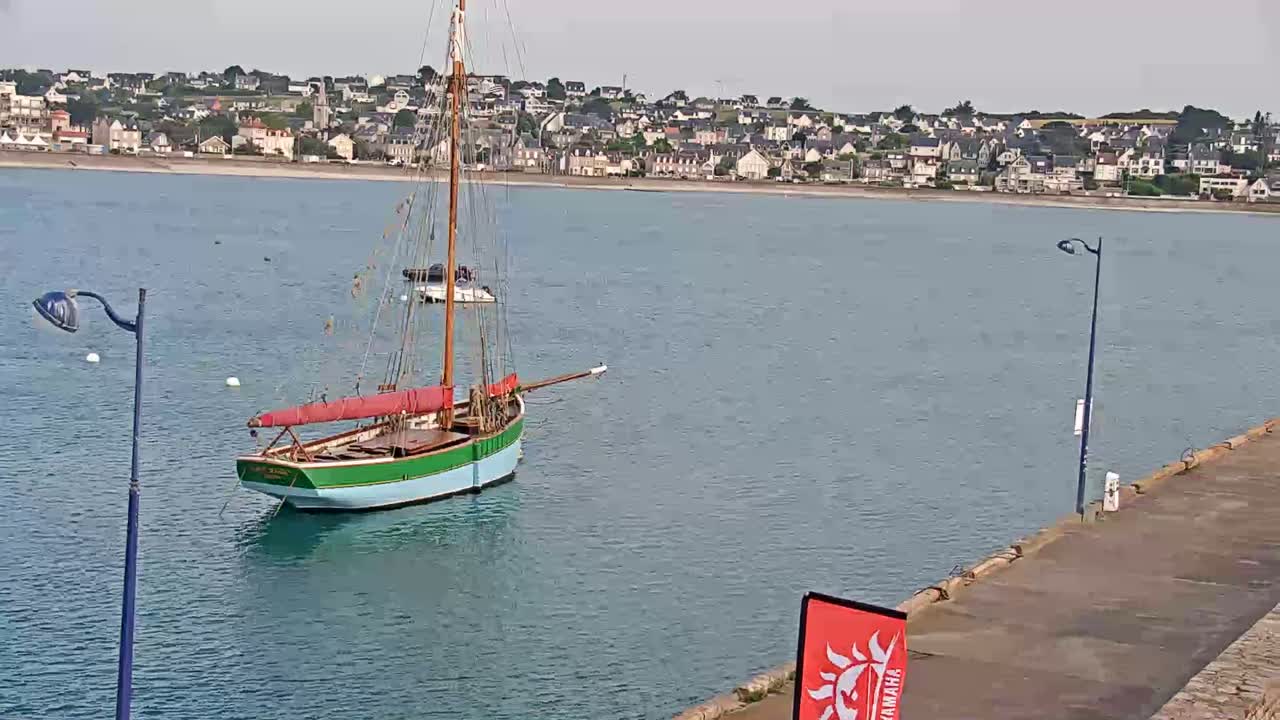 A sailboat is anchored in calm water near a coastal town on a partly cloudy day.
