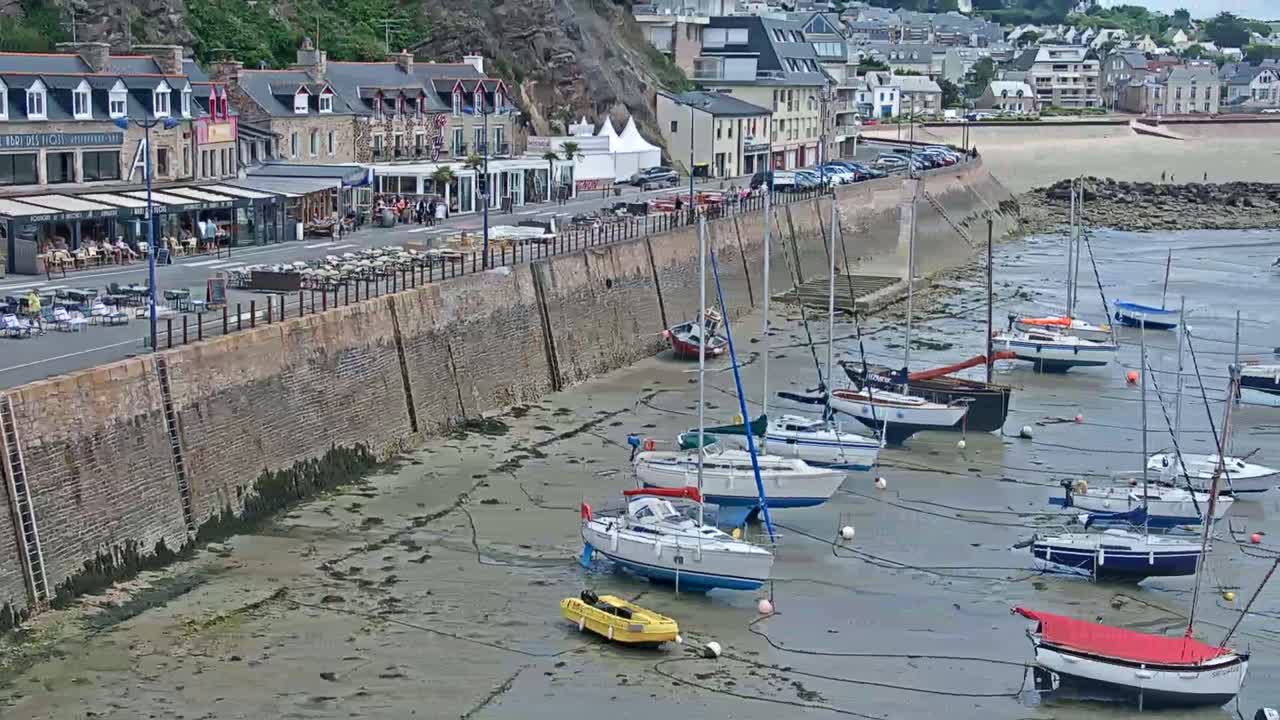 A waterfront town with a stone wall along the shore, numerous sailboats grounded on a tidal flat, and buildings and restaurants behind the wall.