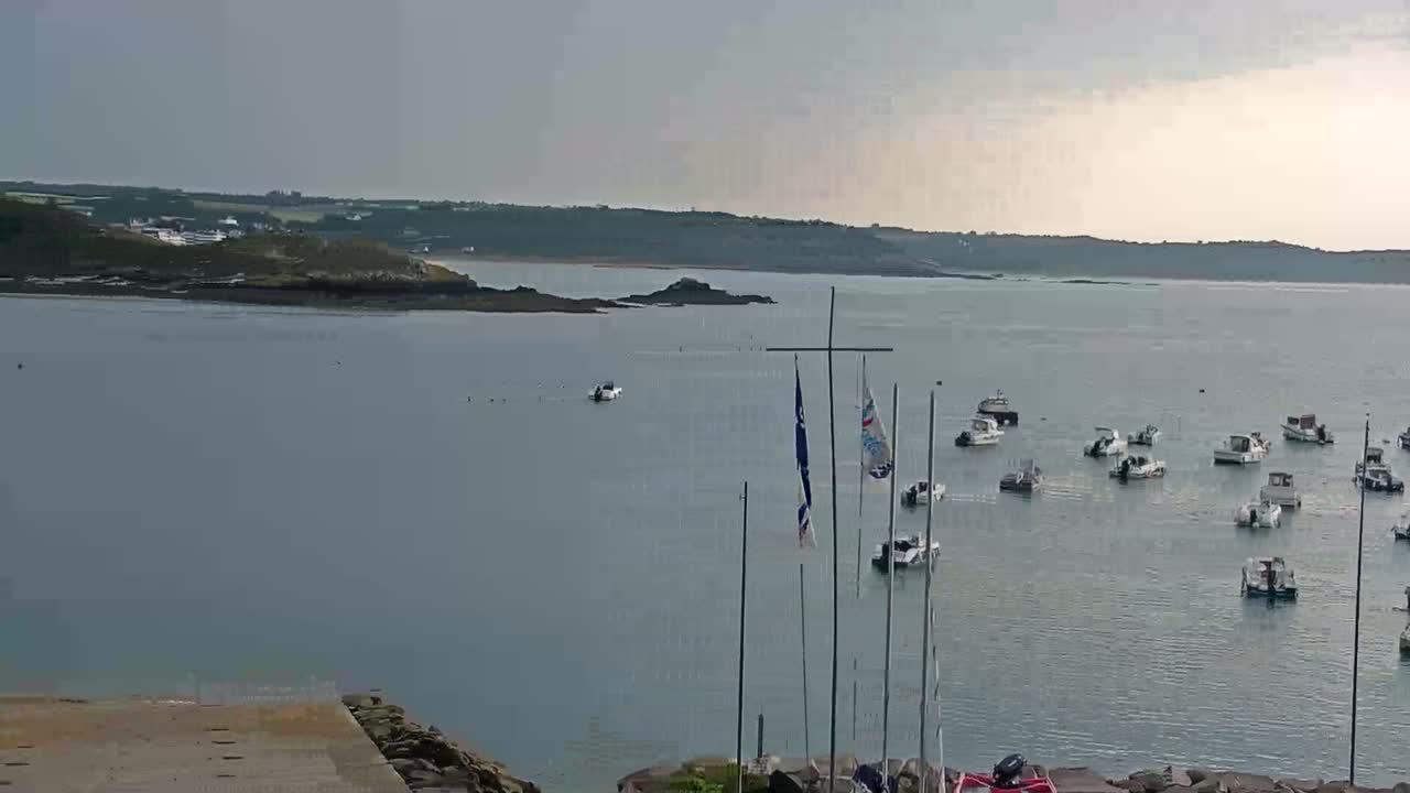 A calm, overcast day shows numerous boats moored in a bay, with a rocky shoreline and distant land visible in the background.