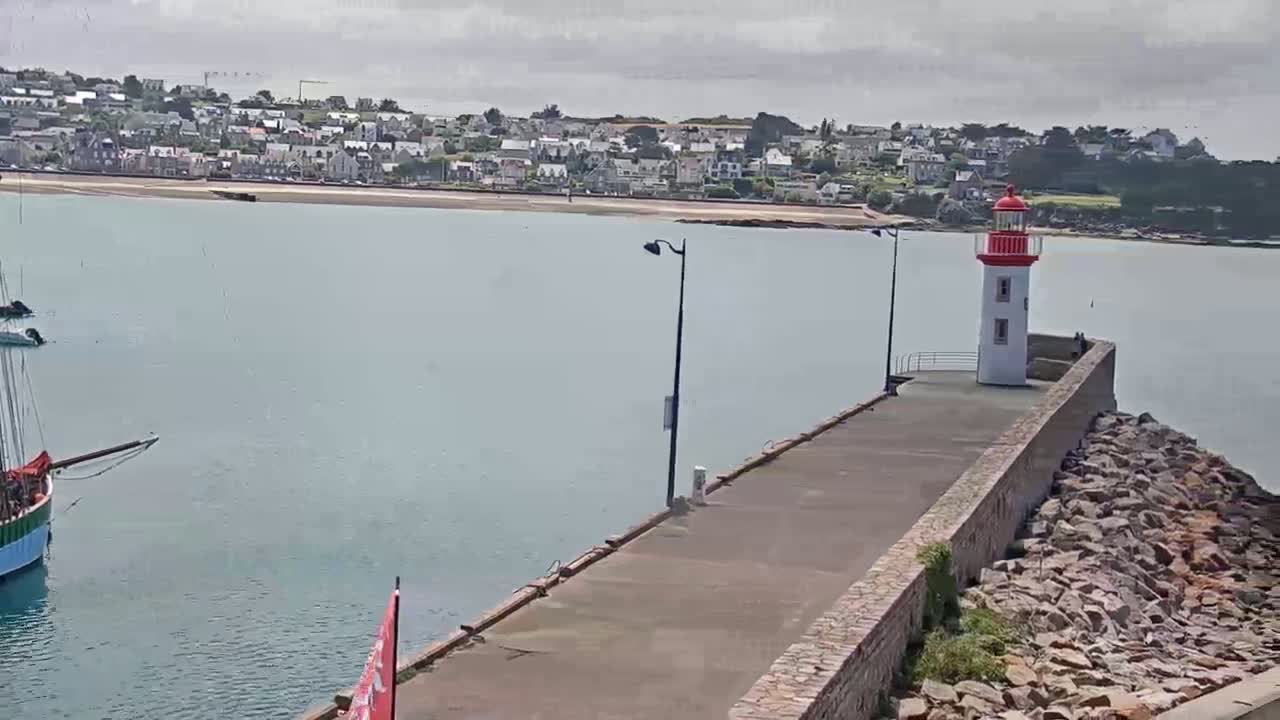 A red and white lighthouse sits at the end of a long pier extending into a calm body of water, with a coastal town visible in the background under an overcast sky.