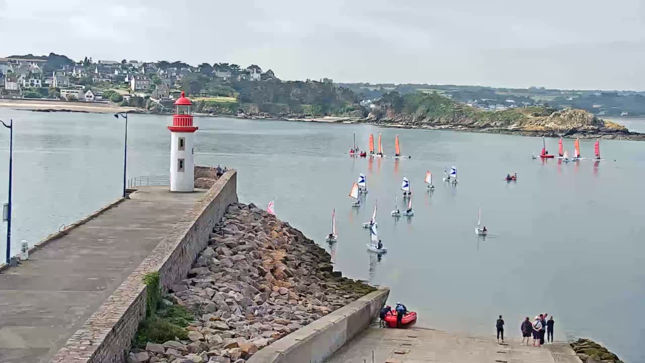 A red and white lighthouse stands on a stone jetty overlooking a calm body of water with numerous sailboats and a few people gathered on the shore.