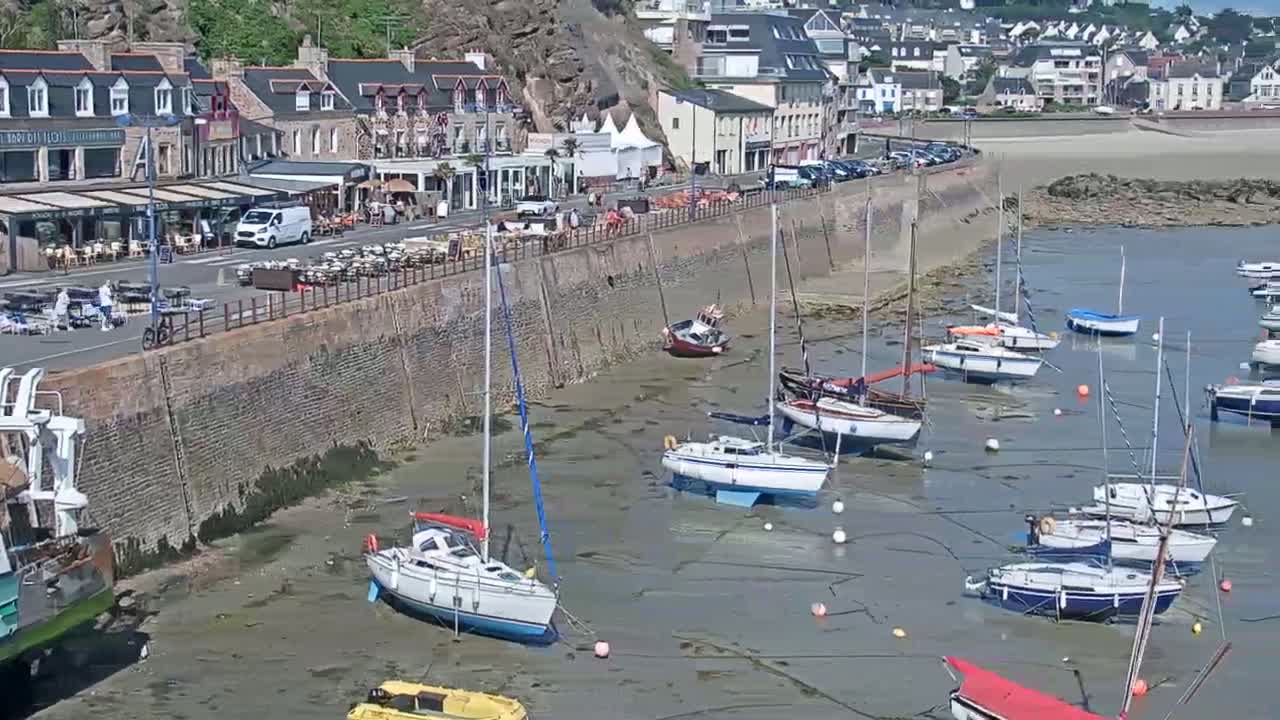 Many sailboats are moored in a harbor at low tide next to a stone wall and a row of buildings.