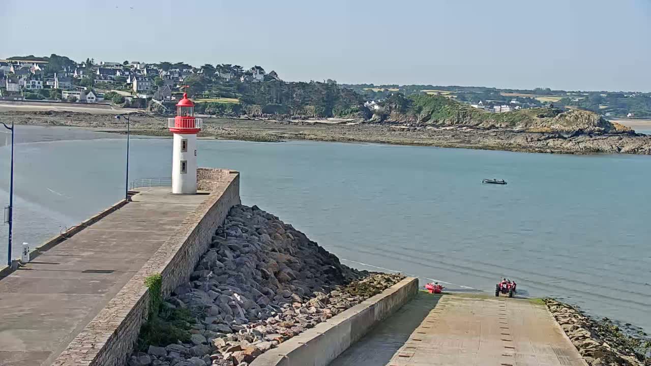 A red and white lighthouse stands on a stone pier overlooking a calm body of water, with a small town and rocky coastline visible in the background under a clear sky.