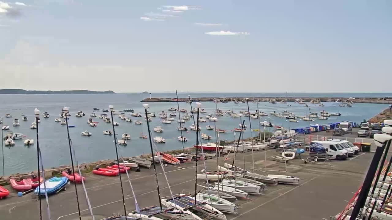 Many sailboats are moored in a harbor on a partly cloudy day, with additional sailboats and other small watercraft docked on land.