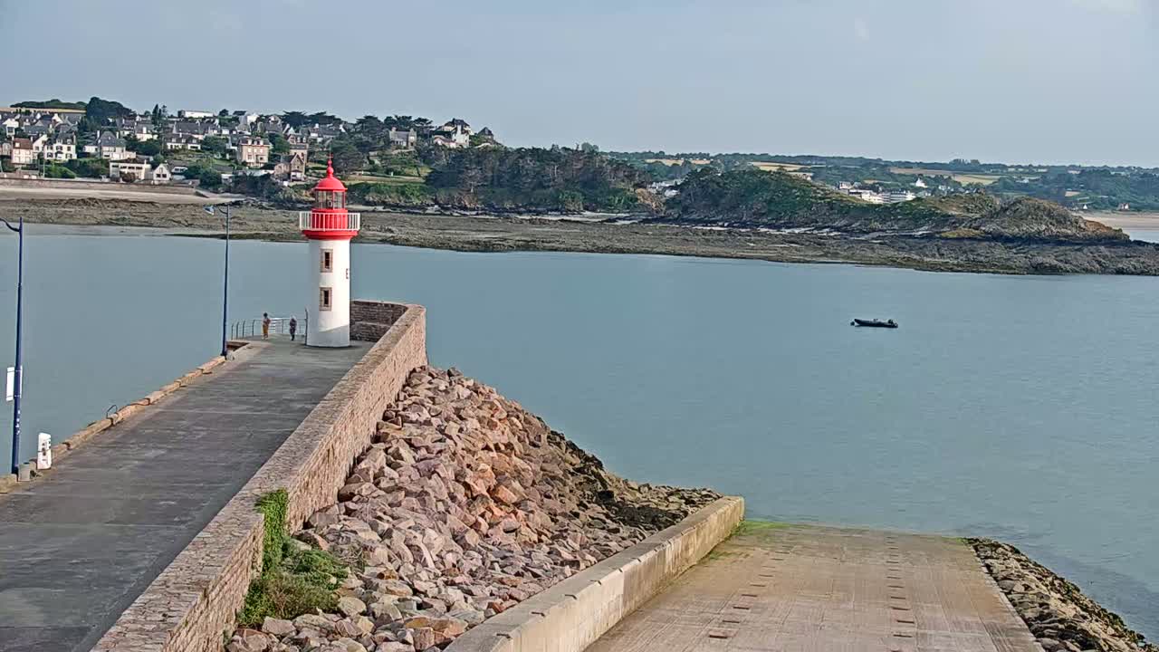 A red and white lighthouse stands on a stone jetty overlooking a calm body of water, a small boat visible in the distance, with a coastal town and low hills in the background on a clear day.