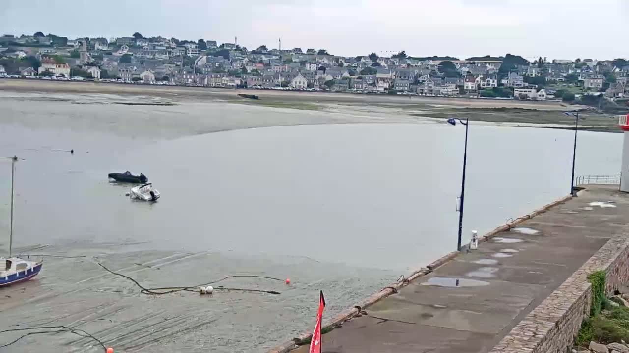 A coastal town is visible in the distance across a calm body of water with low tide exposing a wide expanse of mudflats; two small motorboats are beached nearby, and a small sailboat is moored at a stone pier next to a lighthouse.