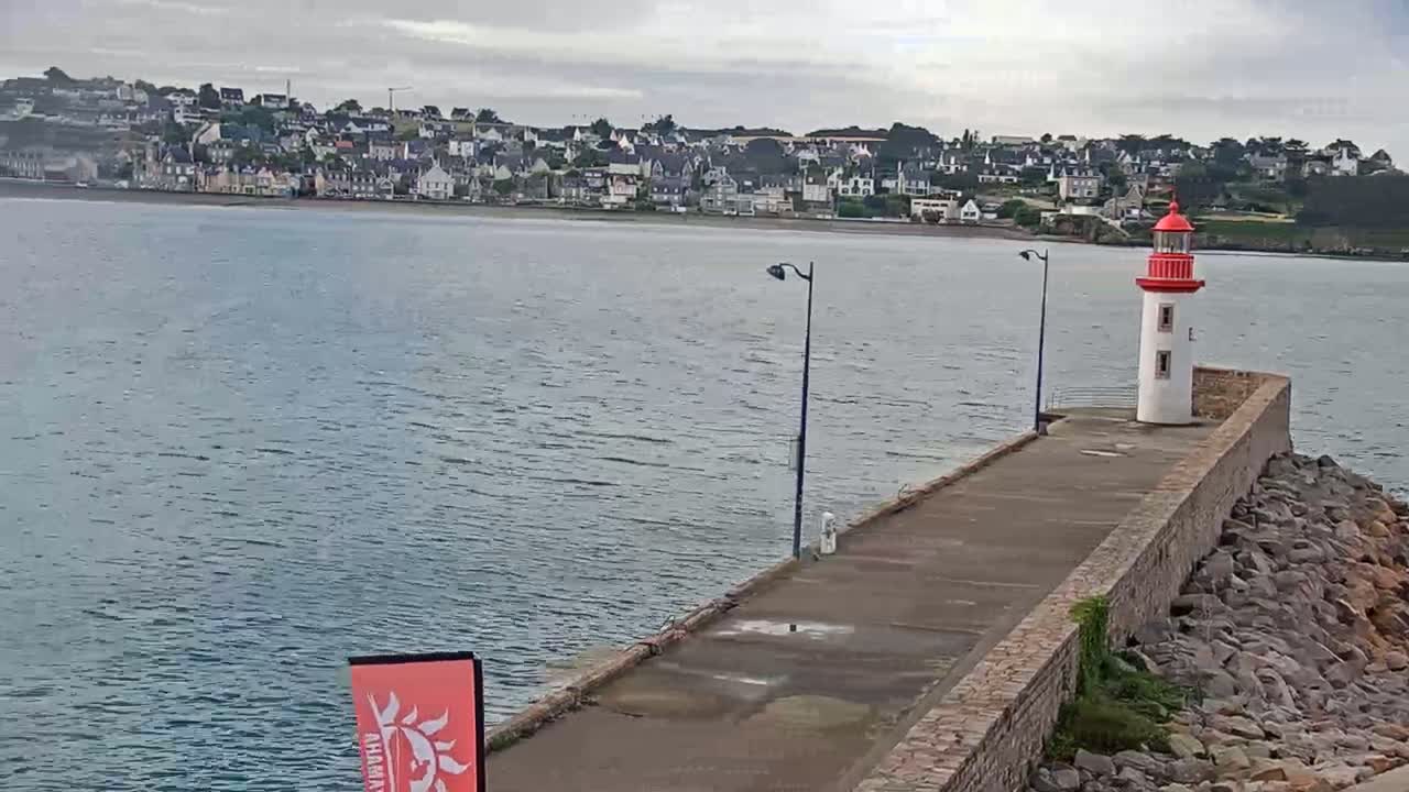 A red and white lighthouse stands at the end of a stone pier overlooking a calm body of water and a town under an overcast sky.