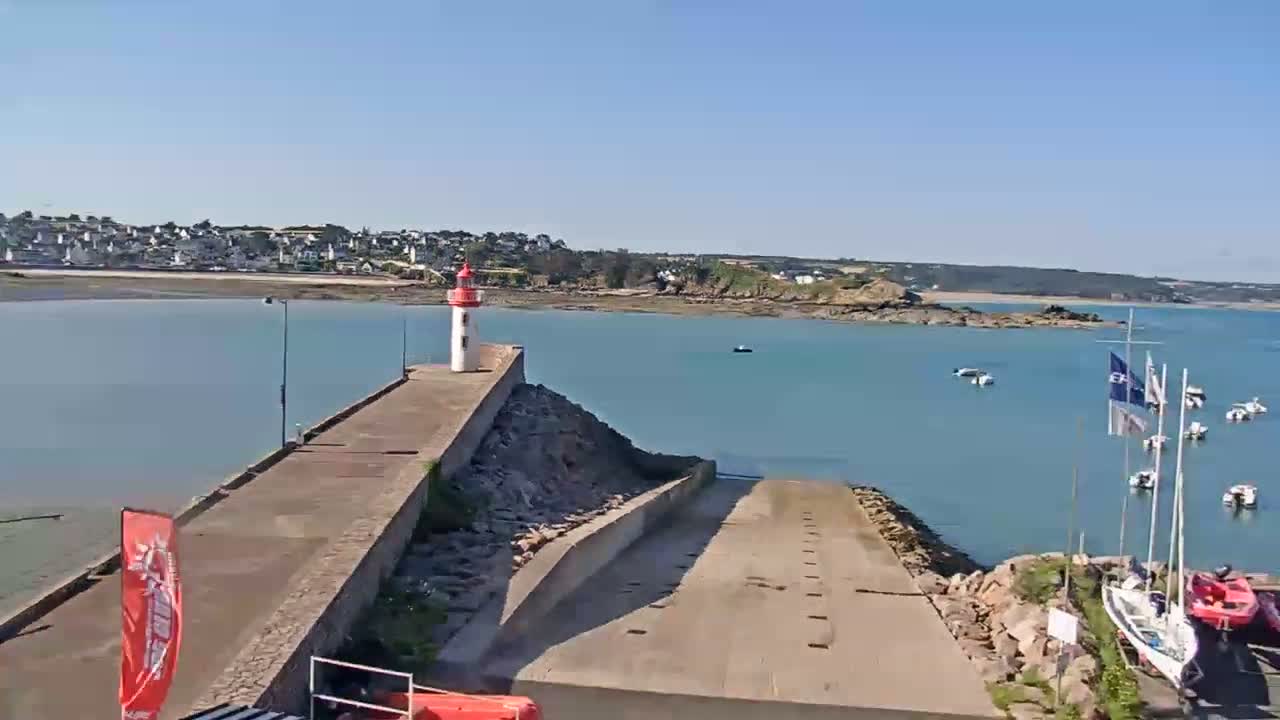 A red and white lighthouse stands on a concrete pier overlooking calm blue water with several sailboats and a small town in the background under a clear, sunny sky.