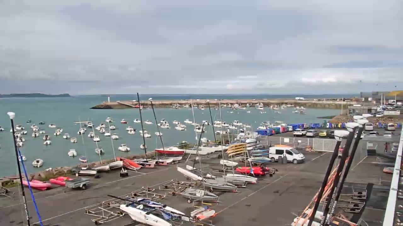 Many boats are moored in a harbor on a cloudy day, with additional boats and trailers stored in a nearby lot.
