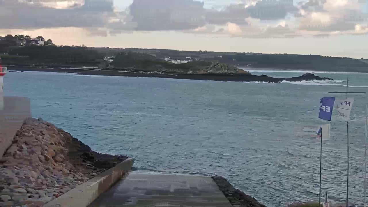 A choppy grey-blue sea meets a concrete boat ramp and rocky shore under an overcast sky, with distant land, houses, and a red-and-white lighthouse on the left, depicting a cloudy coastal day.