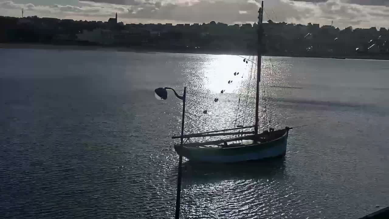 Under a partly cloudy sky with bright sunlight reflecting intensely on the water, a small sailboat and a streetlamp are silhouetted against a distant town skyline.