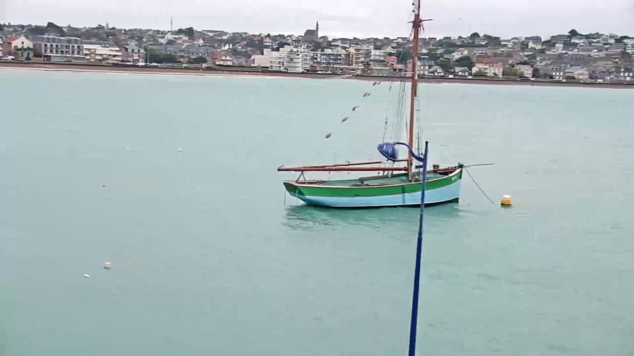 A two-toned green and light blue sailboat is moored in calm, greenish-blue water with a distant coastal town featuring buildings and a church spire along a beach, all under an overcast sky.