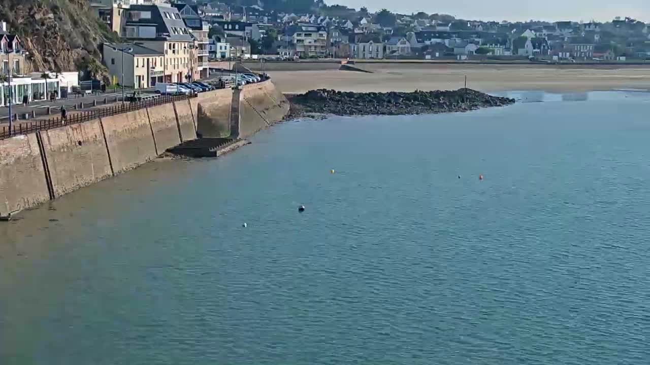 A two-toned green and light blue sailboat is moored in calm, greenish-blue water with a distant coastal town featuring buildings and a church spire along a beach, all under an overcast sky.