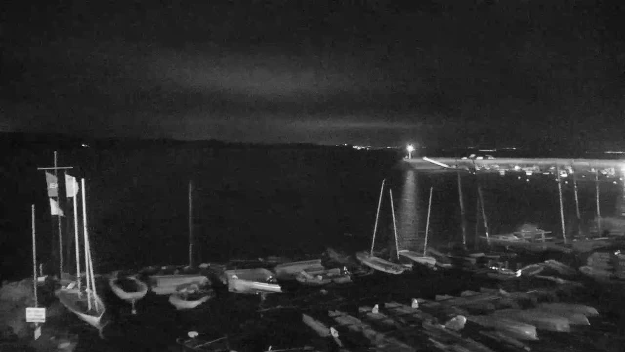 A two-toned green and light blue sailboat is moored in calm, greenish-blue water with a distant coastal town featuring buildings and a church spire along a beach, all under an overcast sky.