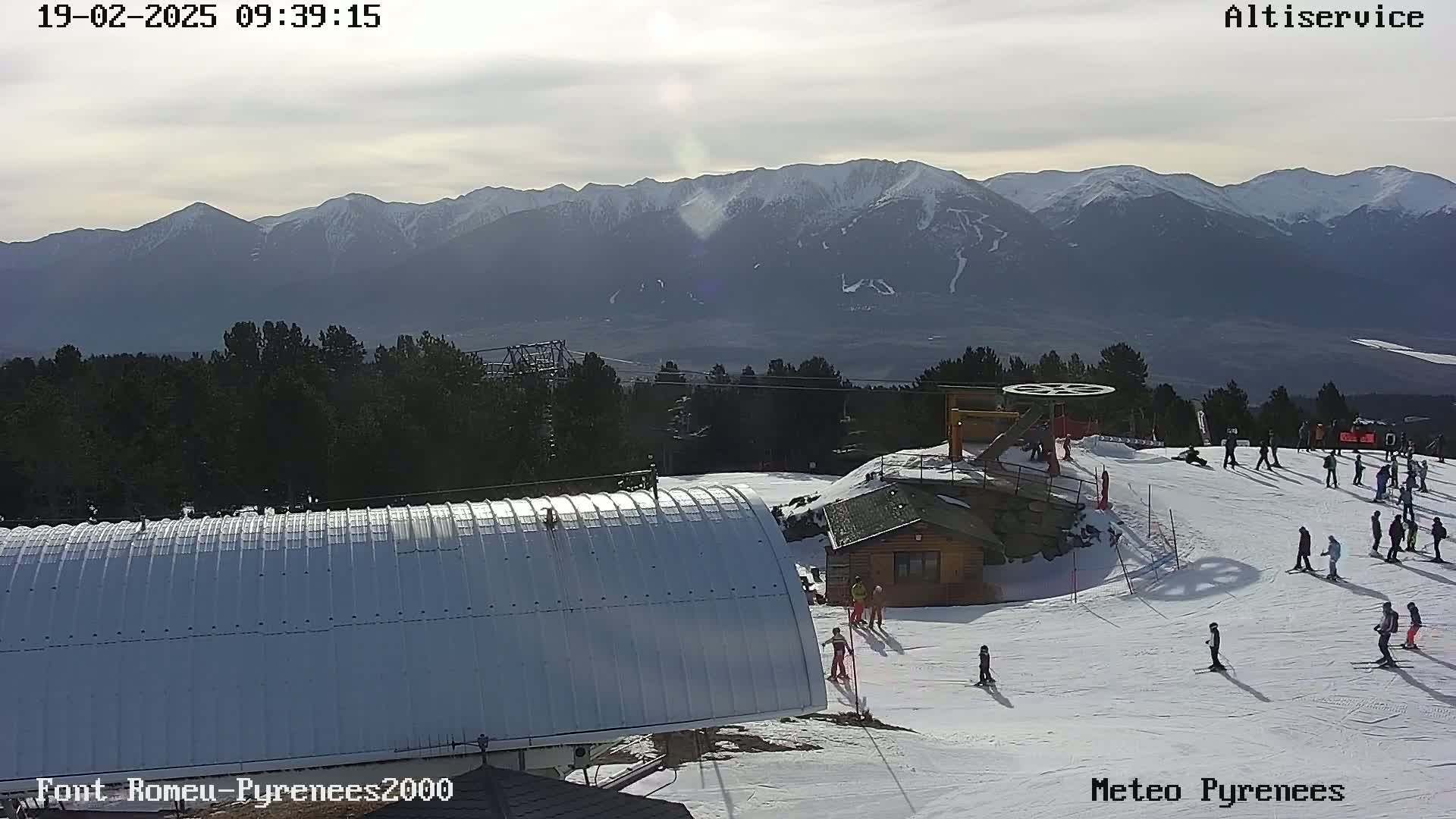 A snowy ski slope with several skiers and a mountain range in the background under a mostly sunny sky.