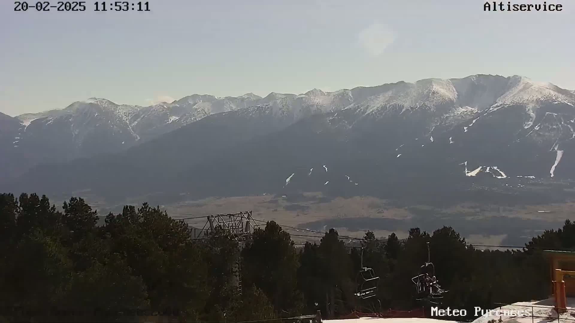 A hazy, sunny day reveals a snow-capped mountain range in the distance, beyond a foreground of dark evergreen trees and ski lift infrastructure.
