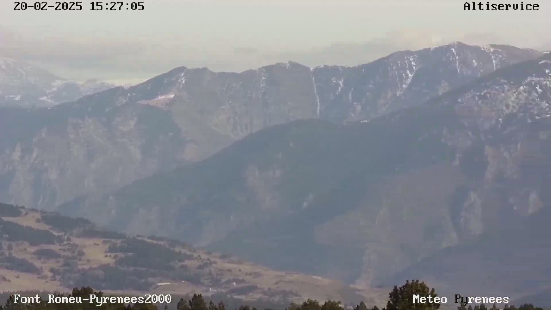 A hazy view shows a vast mountain range with some snow patches on the peaks under a mostly clear sky.