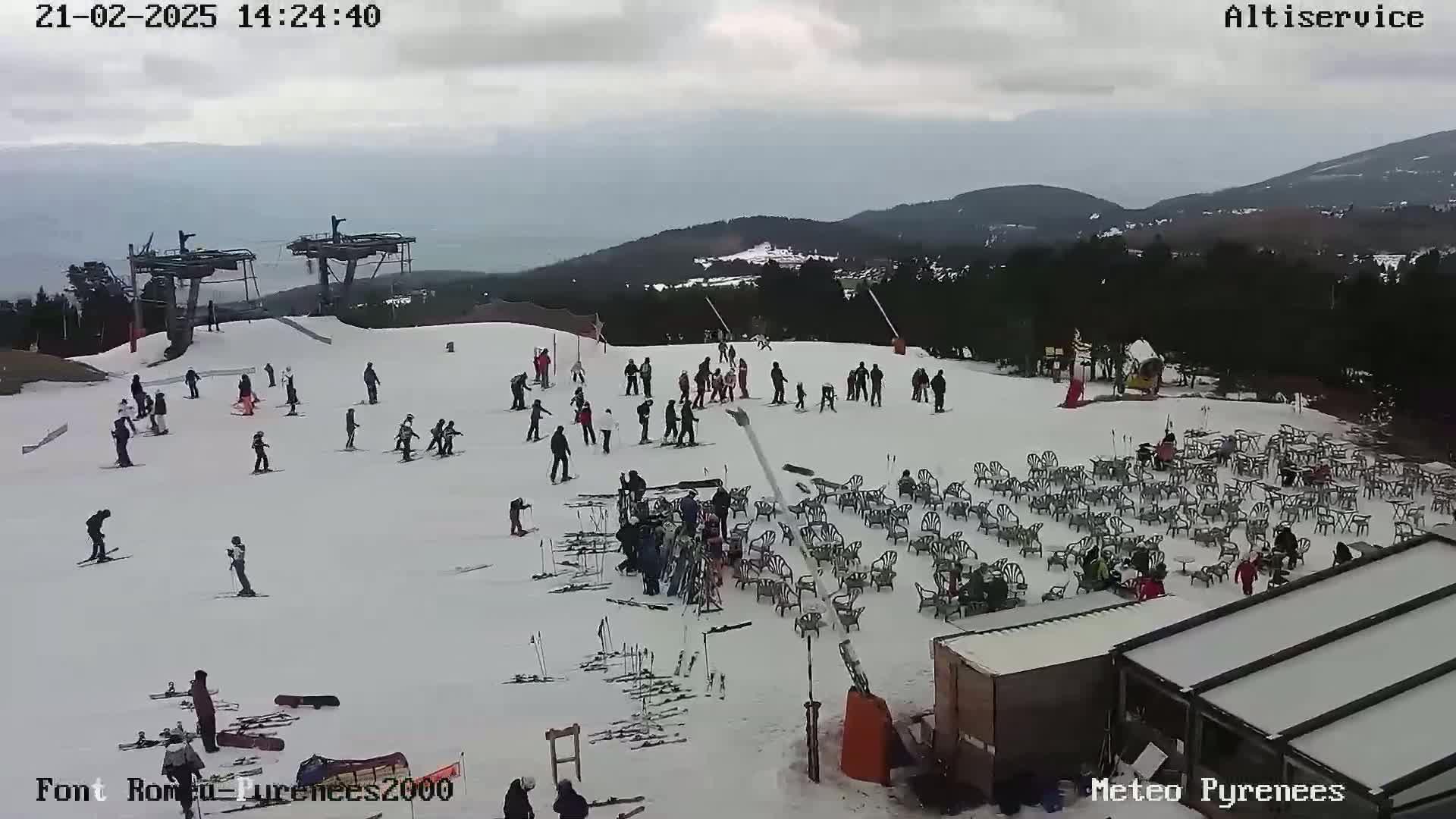 A snow-covered ski slope with skiers and snowboarders, a chairlift, numerous outdoor chairs and tables, and a building in the background under an overcast sky.