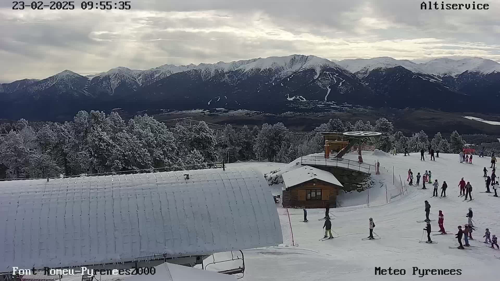 A snow-covered ski slope with skiers and snowboarders, a small building, and a chairlift, overlooks a valley and a snow-capped mountain range under a partly cloudy sky.