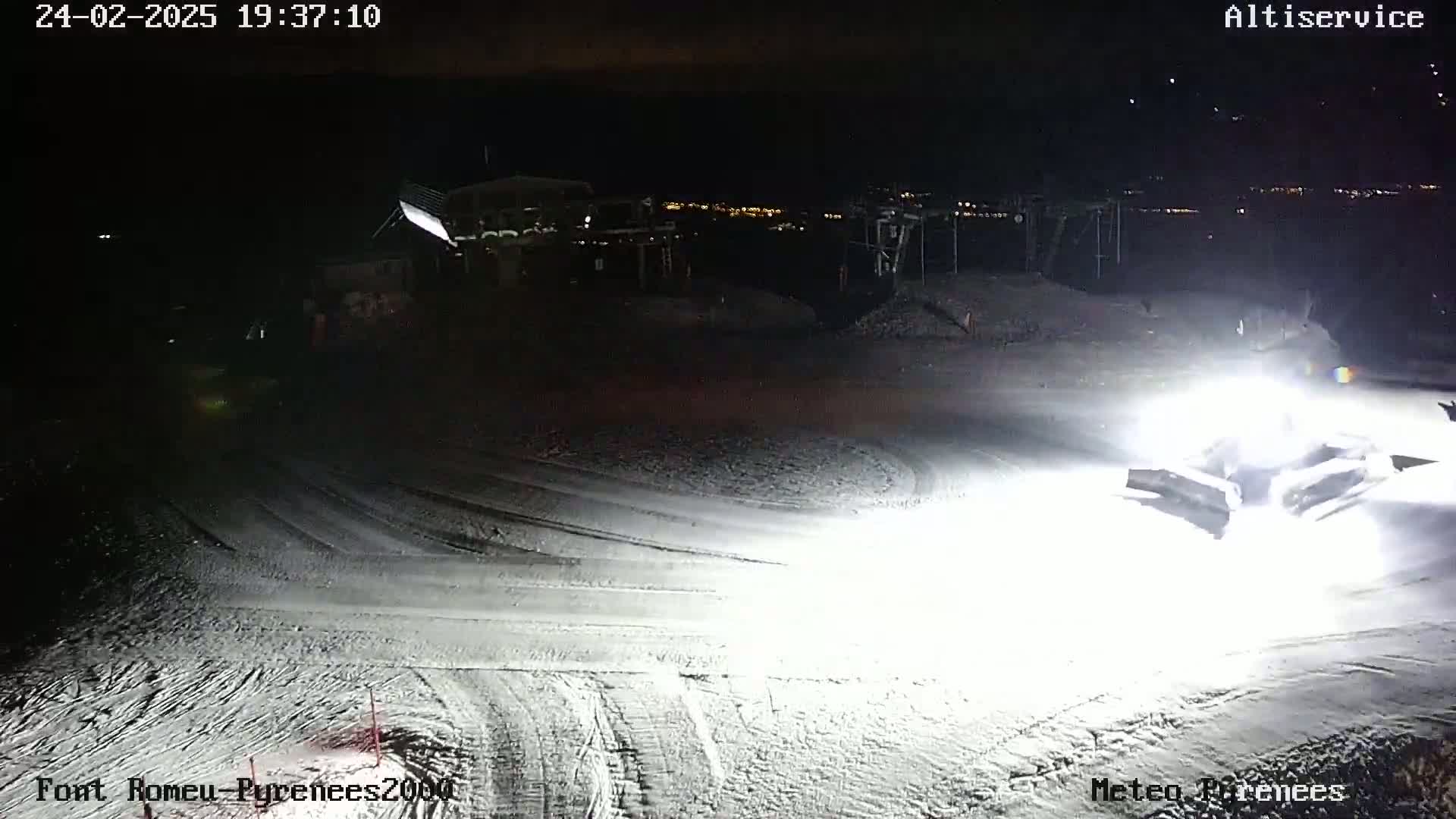 A snow groomer illuminates a snow-covered slope at night, with distant lights visible in the background.