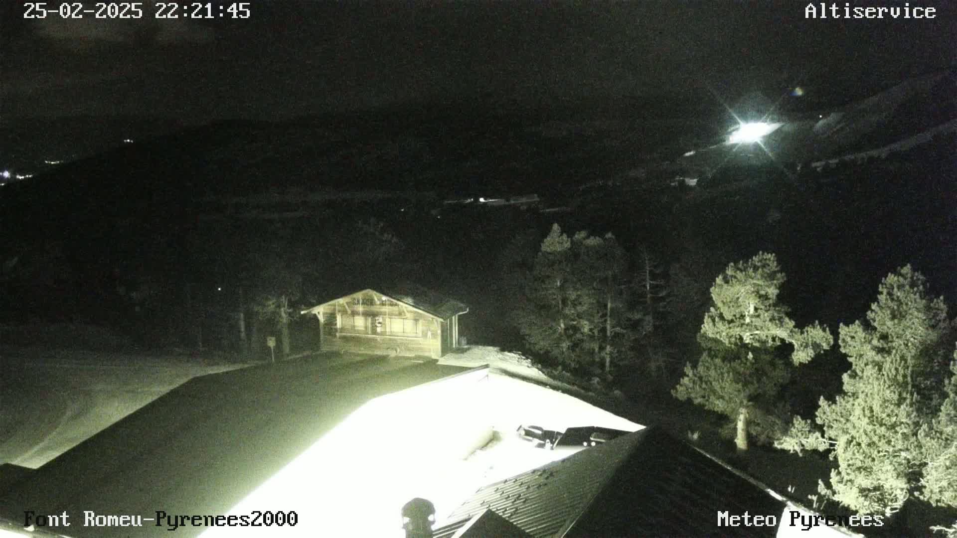 A nighttime, snow-covered landscape with a small building illuminated, surrounded by dark evergreens under a clear sky.