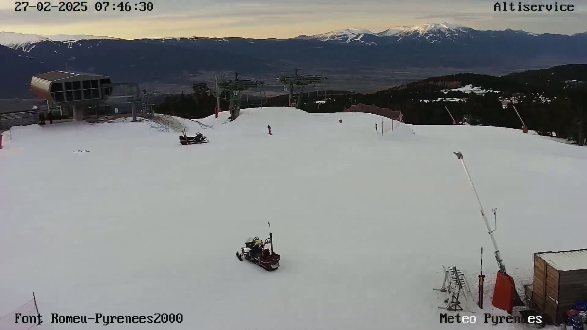 A snow-covered ski slope with snowmobiles, ski lifts, and a mountain range in the background under a cloudy sky.