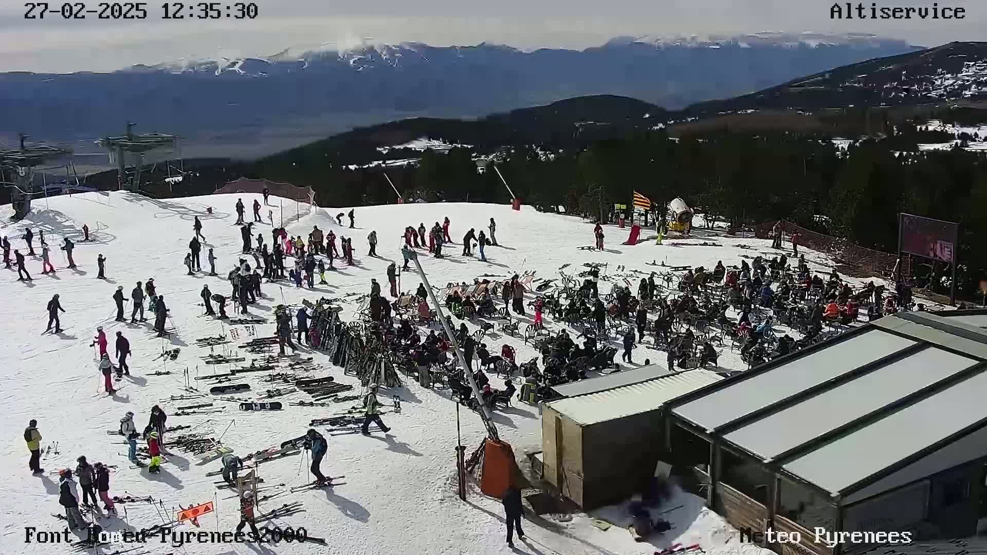 A sunny day at a snow-covered ski resort shows numerous skiers and snowboarders, along with many people seated at outdoor tables near ski equipment and lift chairs, with mountains in the background.