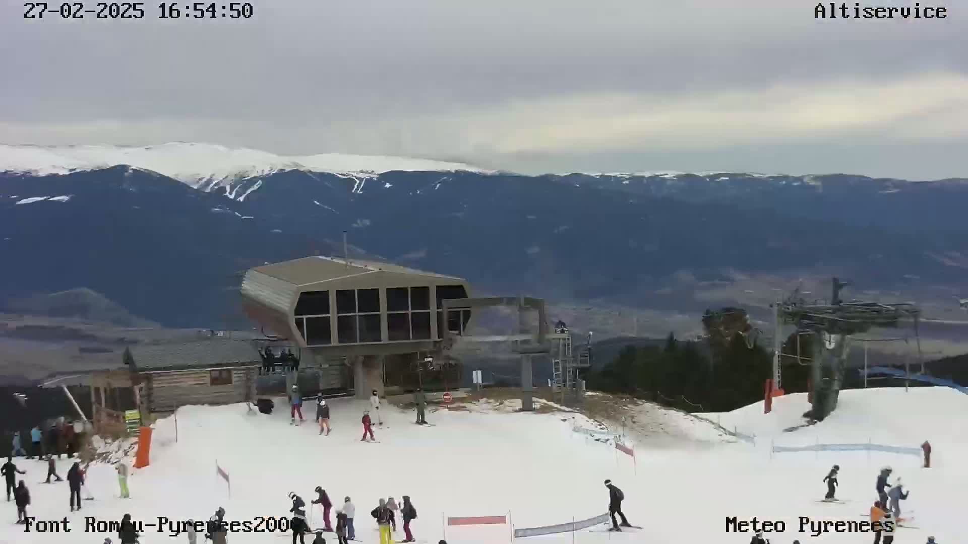 A ski lift station with skiers and snowboarders on a snowy mountain slope under an overcast sky.