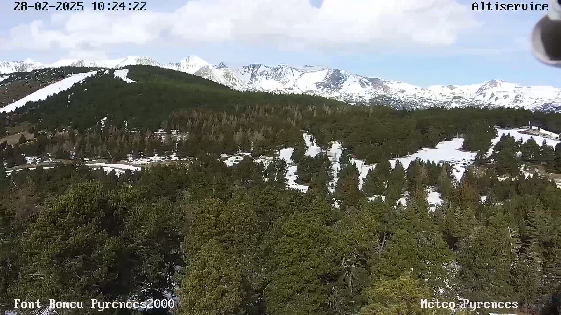 A partly cloudy day shows a mountainous landscape with snow-capped peaks in the background and a dense evergreen forest in the foreground, speckled with patches of snow.