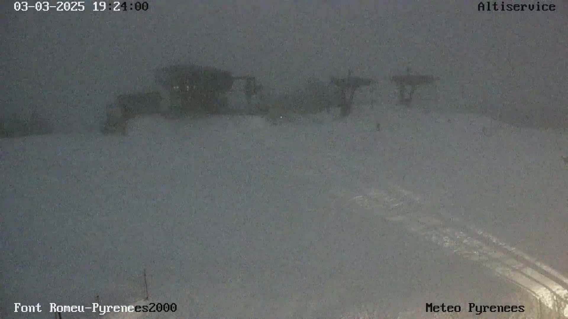 A snow-covered landscape at night shows indistinct structures in the distance under a dark, overcast sky.
