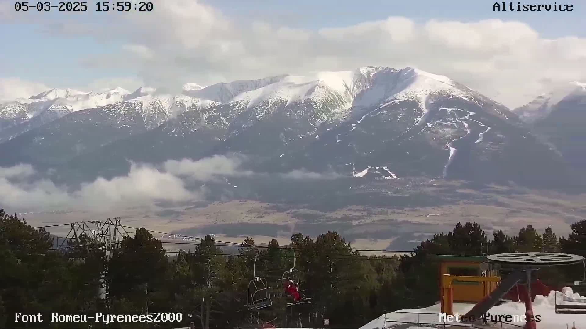 A partly cloudy day reveals snow-capped mountains in the distance, with a chairlift and skiers visible in the foreground.