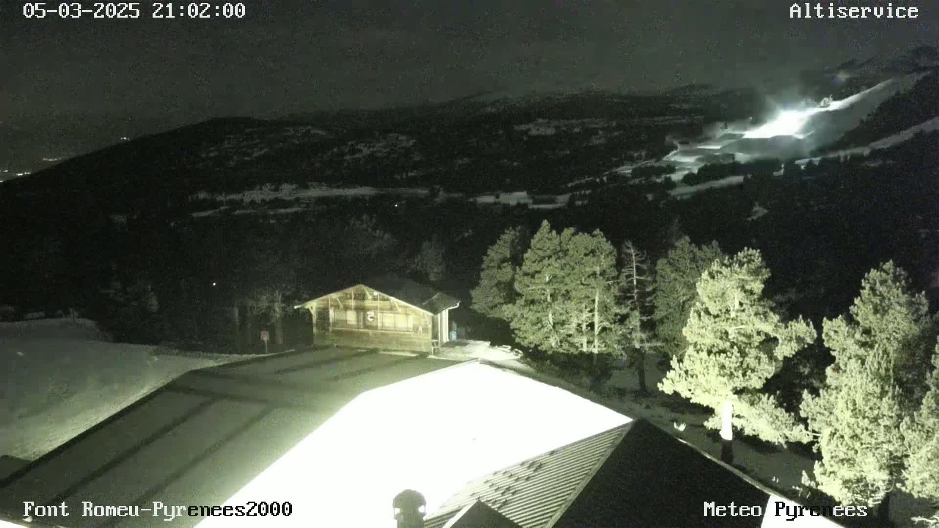 A nighttime view of a snow-covered mountain landscape, illuminated by artificial lights, shows a building and several evergreen trees.