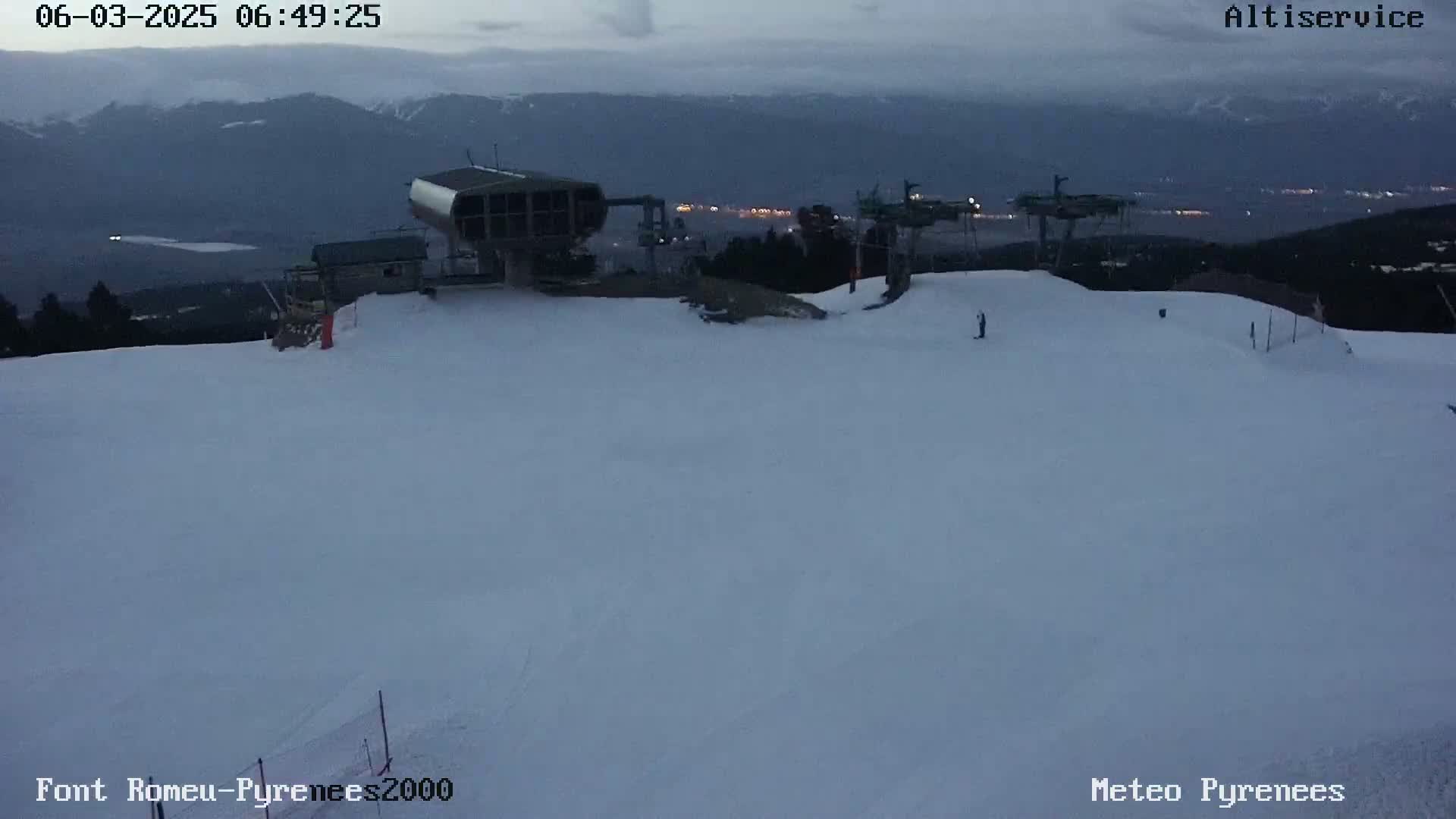 A snow-covered ski slope at dusk, with ski lifts and a distant mountain range visible under a cloudy sky.