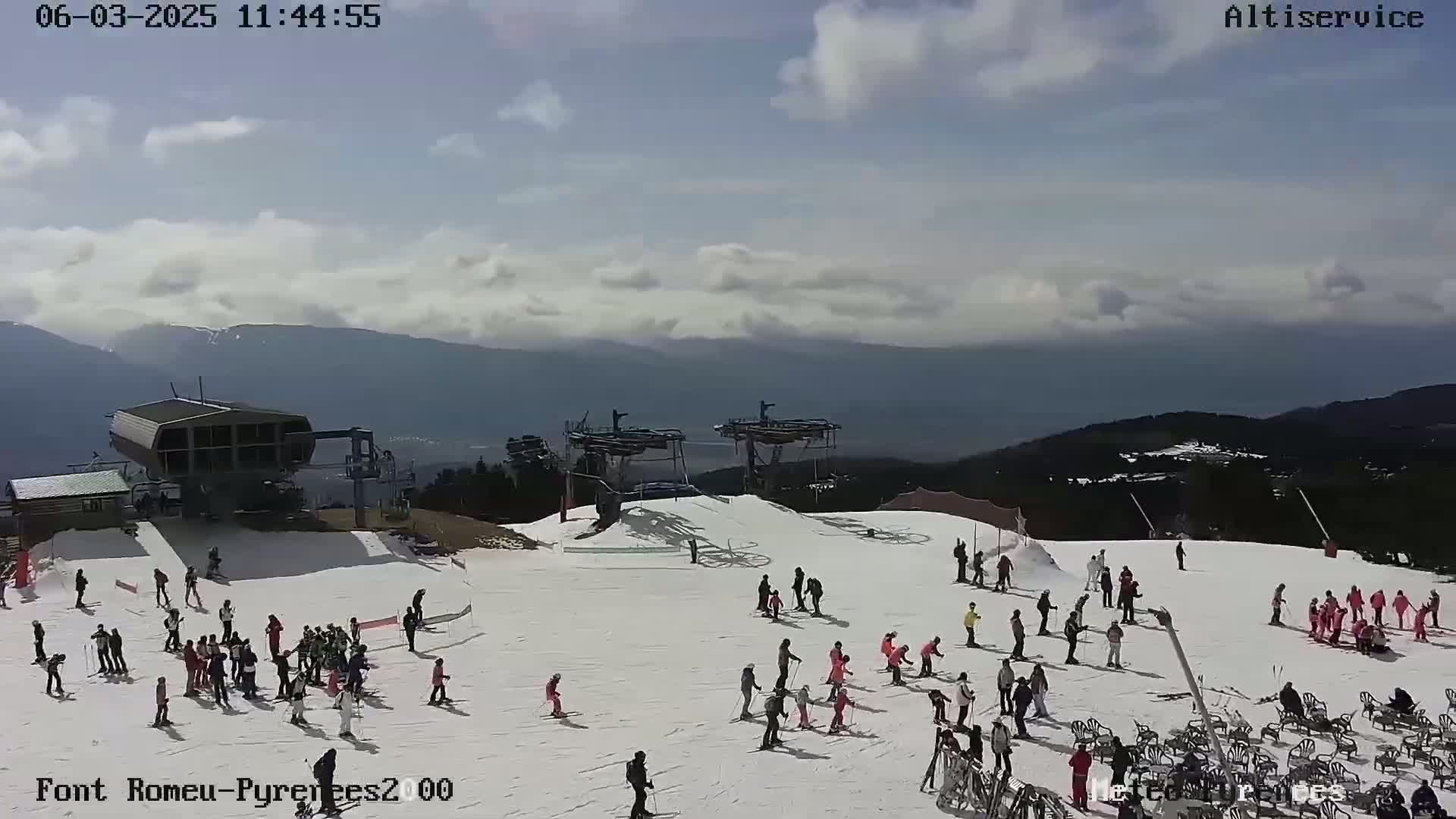 A snowy ski slope with several people skiing and standing around, ski lifts, and a mountain range in the background under a partly cloudy sky.
