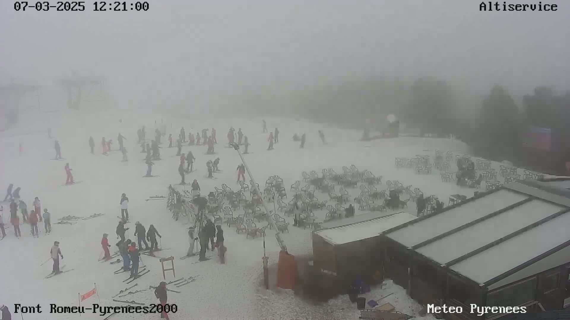 A snowy ski slope is visible through a thick fog, with numerous skiers scattered across the area and many empty chairs set up near a building.
