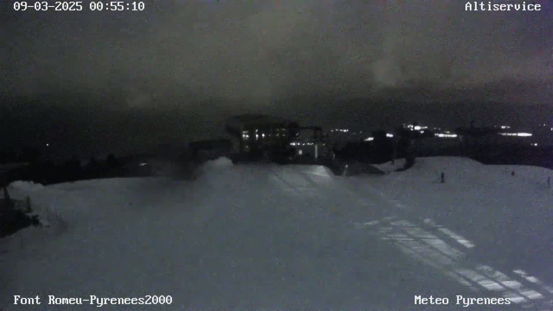 A nighttime view of a snow-covered slope with a building and distant lights visible under a dark sky.