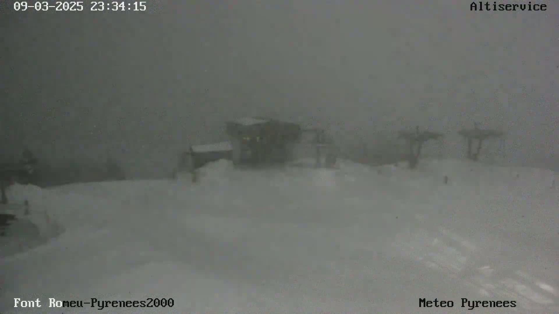 A snow-covered landscape with a building and ski lift towers is barely visible through heavy snowfall.