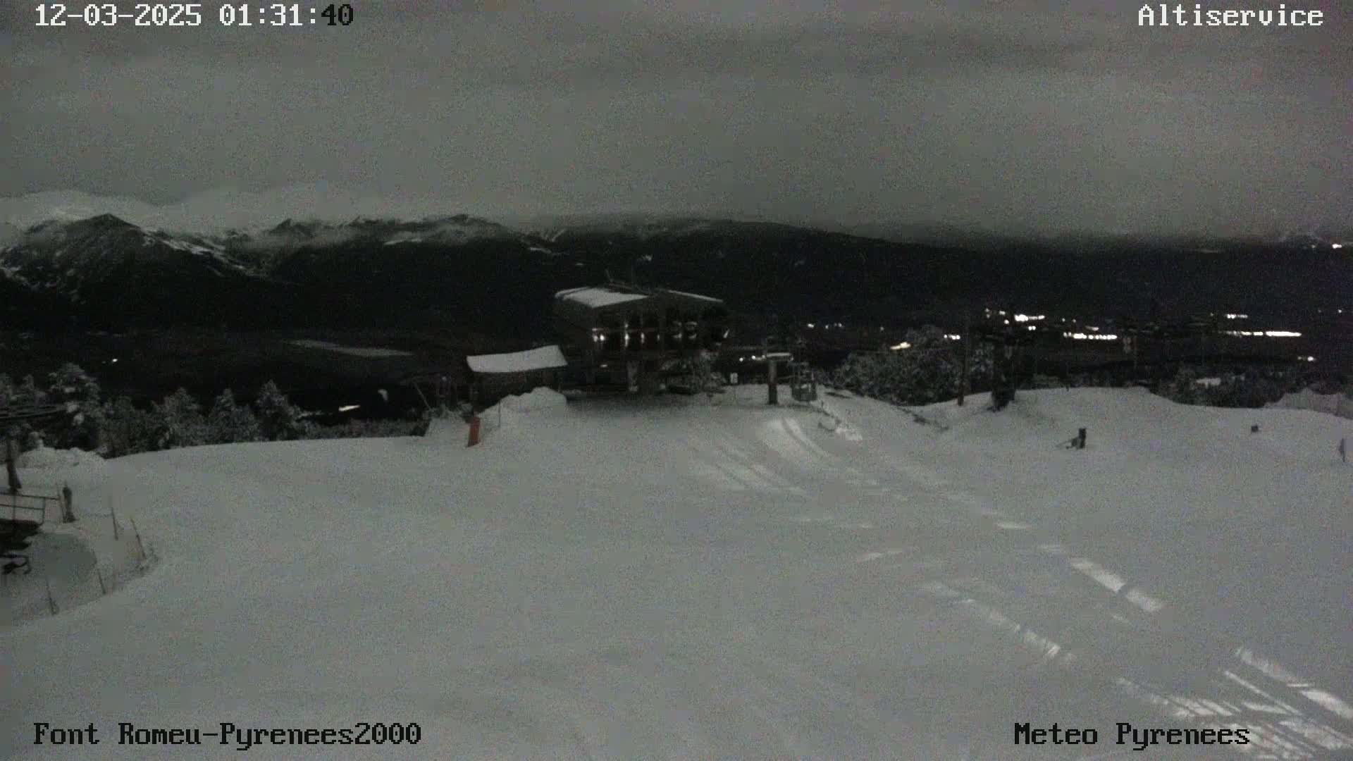 A snow-covered ski slope at night, under a dark, overcast sky, features a building with lights, and distant mountain peaks visible in the background.