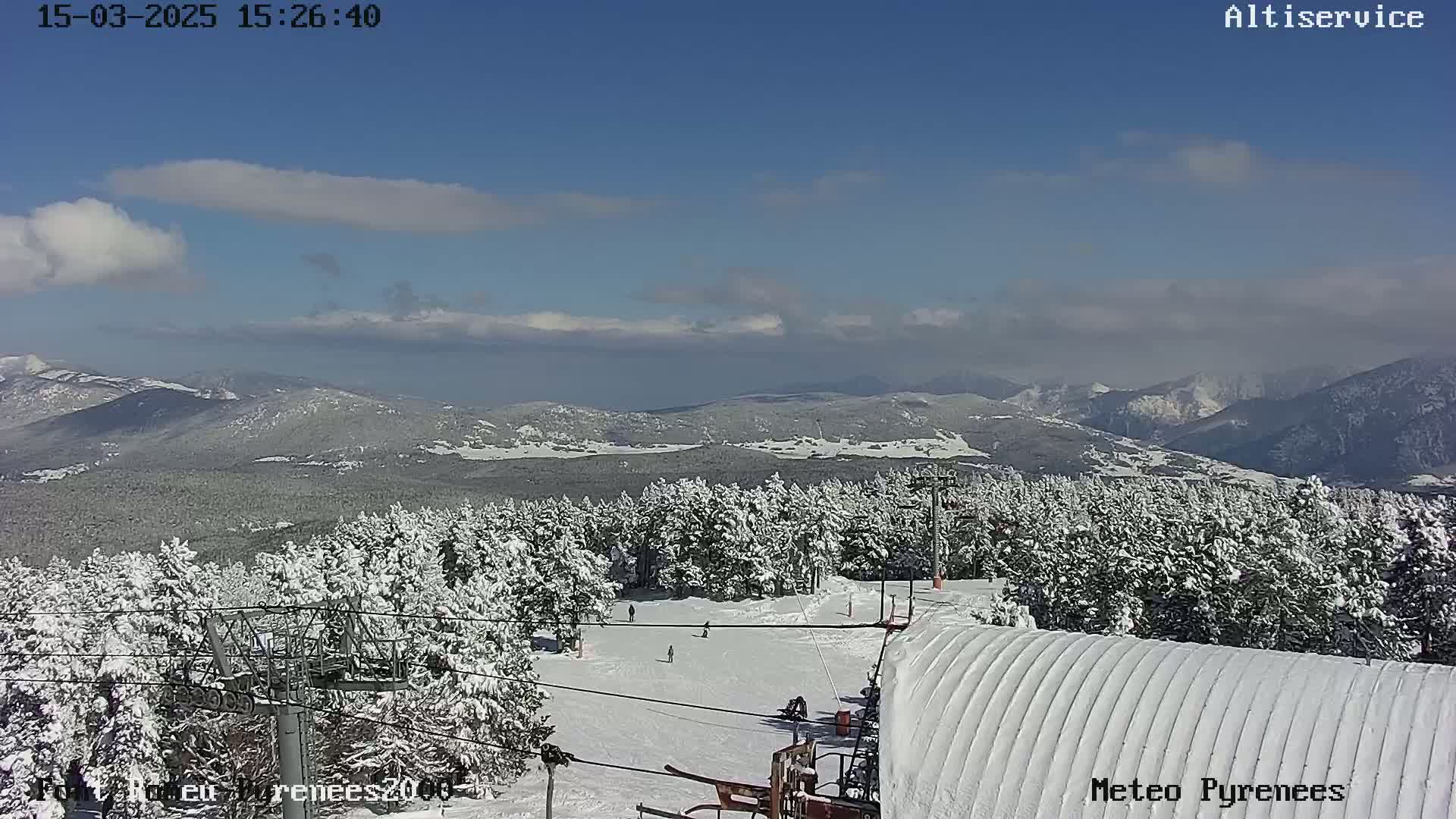 A snow-covered ski slope with several skiers is visible, overlooking a snow-covered forest and distant mountains under a mostly clear, sunny sky.