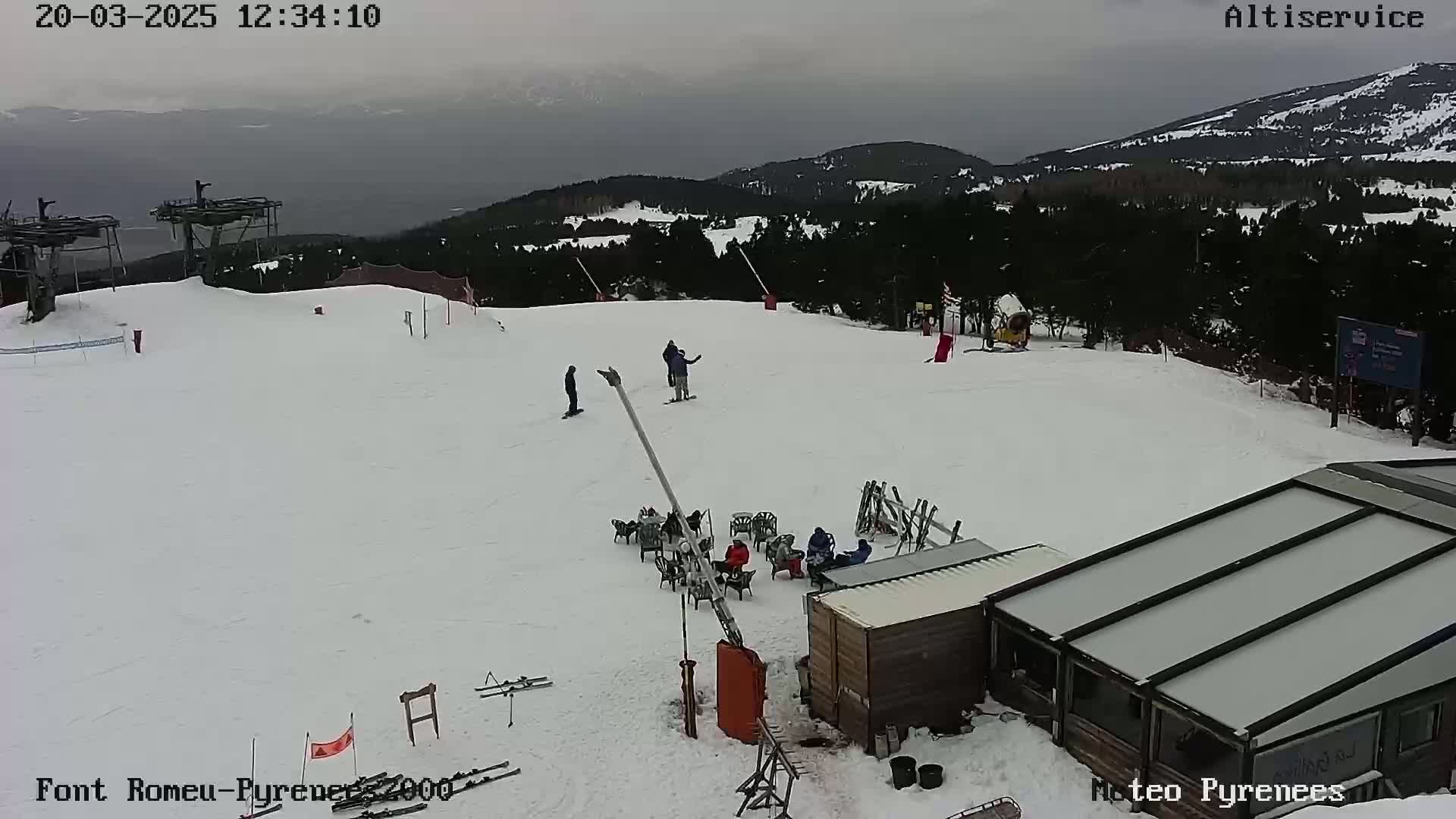 A snow-covered ski slope with several skiers and snowboarders, a small building, and ski lift infrastructure under an overcast sky.