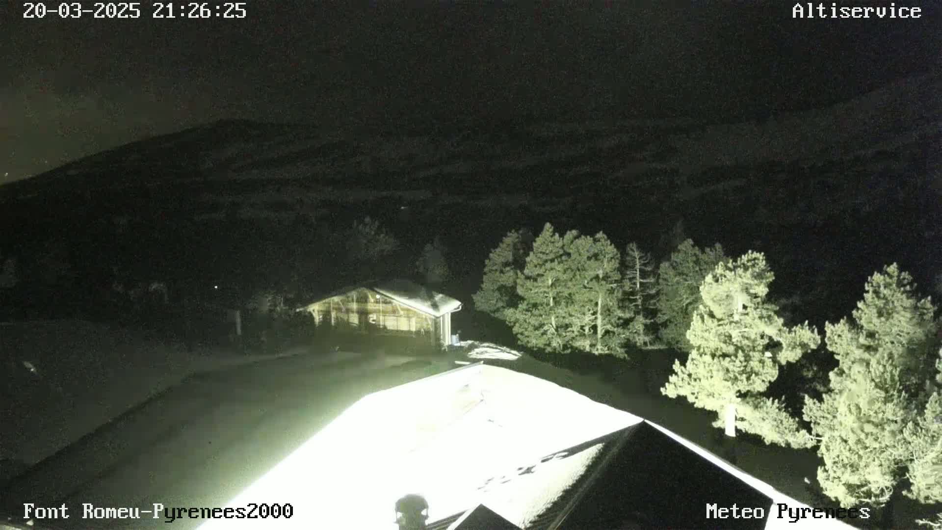 A nighttime, snow-covered scene shows a building illuminated by a bright light, surrounded by dark evergreen trees against a backdrop of a dark mountain under a clear sky.